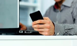 Close-up of male hand holding smartphone and typing on a laptop keyboard