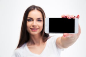 Happy young woman showing blank smartphone screen isolated on a white background. Looking at camera. Focus on smartphone
